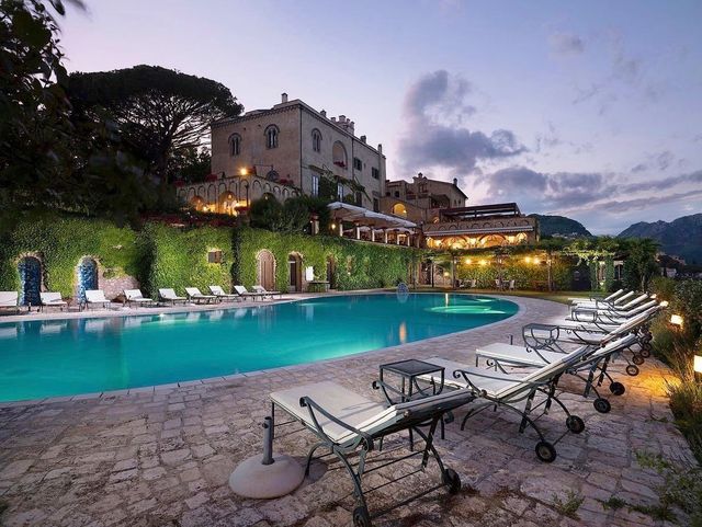 Piscina serale del resort con lettini, villa ricoperta di edera e cielo nuvoloso in riva all'acqua.