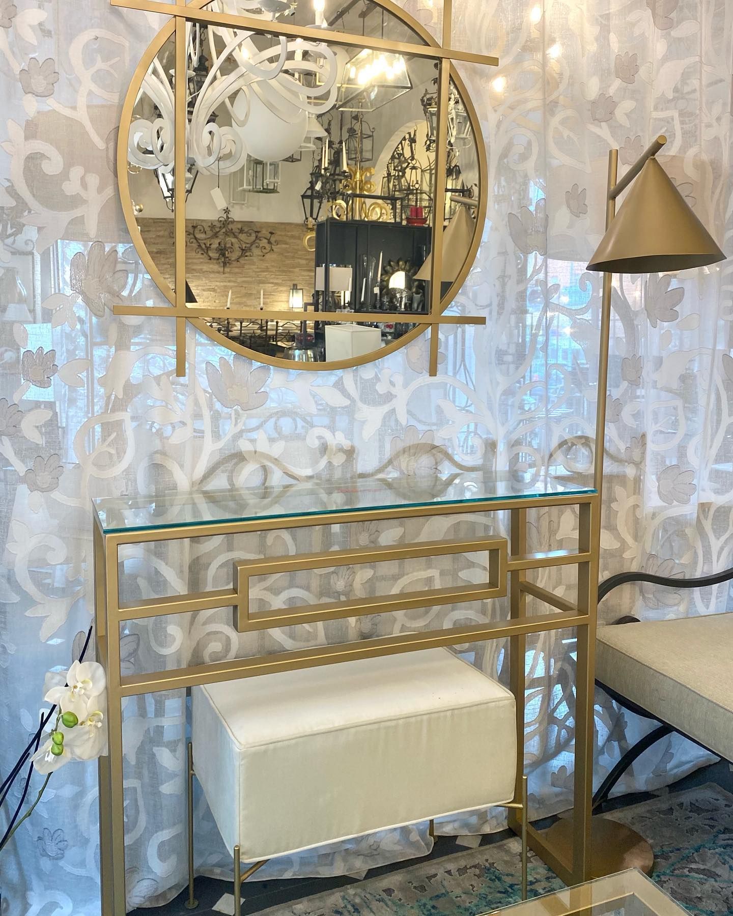 Consolle con cornice dorata e specchio, pouf color crema e lampada da terra, il tutto su uno sfondo di carta da parati floreale.
