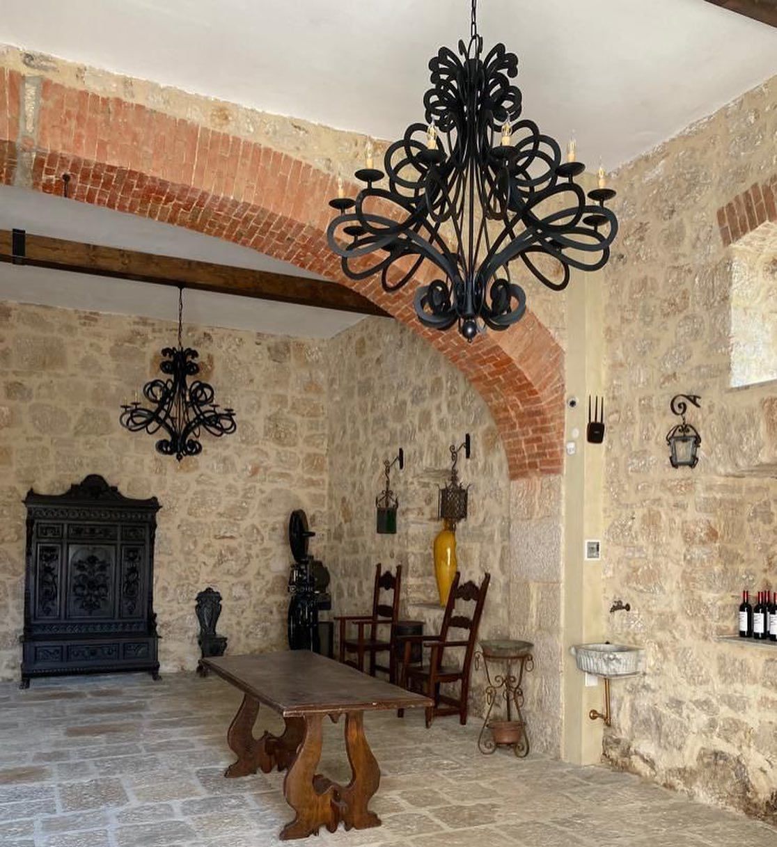 Stanza rustica con pareti in pietra, tavolo e sedie in legno, lampadario nero decorato e armadio antico.