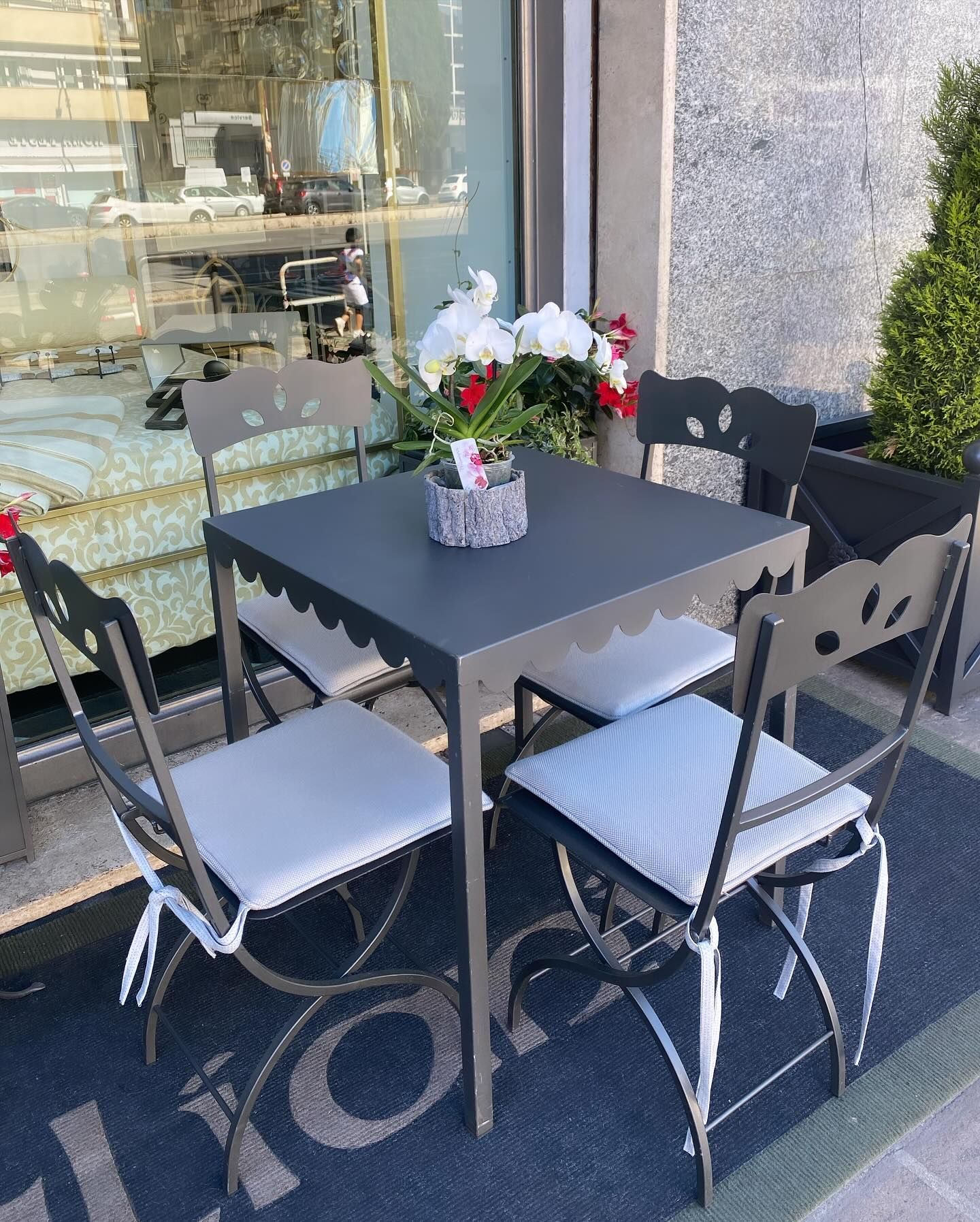 Tavolino da caffè all'aperto con quattro sedie, fiori bianchi in un vaso, accanto alla vetrina di un negozio.