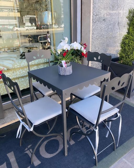 Tavolino da caffè all'aperto con quattro sedie, un centrotavola floreale e la vetrina di un negozio sullo sfondo.