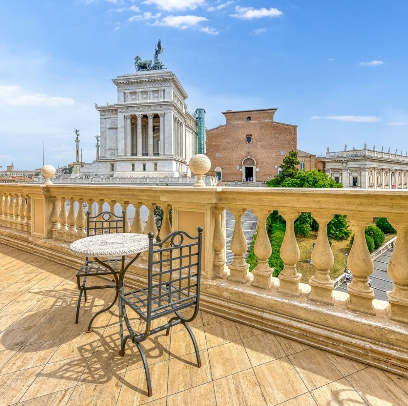 Terrazza in pietra con balaustra decorata e sedia, affacciata su edifici storici sotto un cielo azzurro brillante.