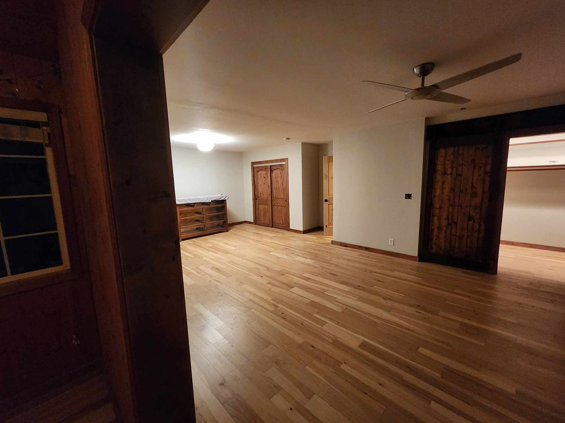Empty room with hardwood floors, white walls, and wooden doors, lit by bright ceiling light and a ceiling fan.