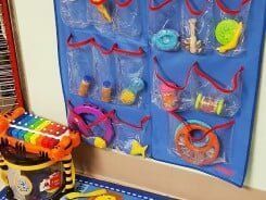 A blue hanging organizer with clear pockets holding various colorful baby toys on a wall.