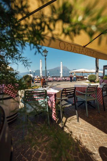 Patio del ristorante sull'acqua con tavoli, ombrelloni, fiori e montagne sullo sfondo.