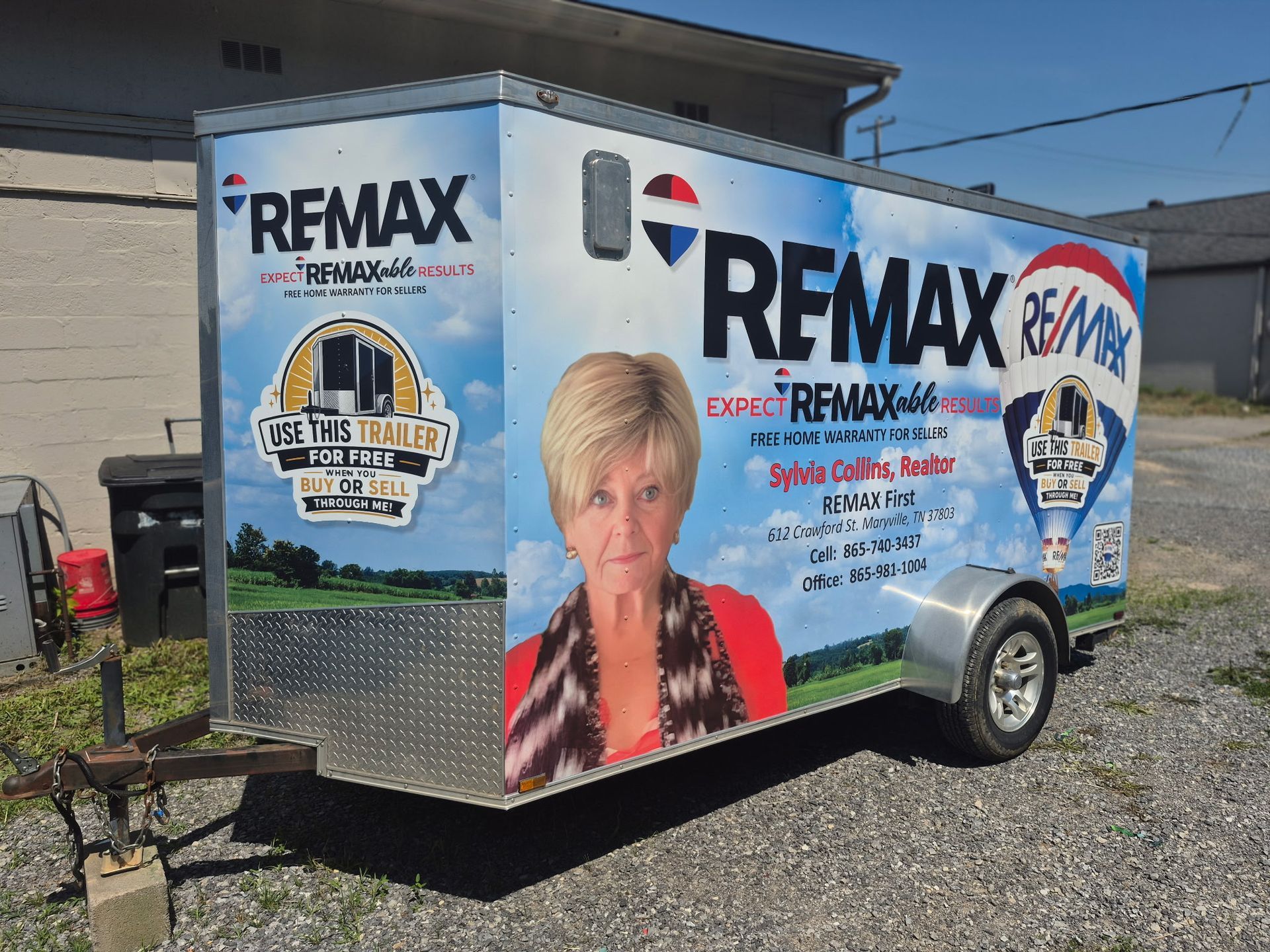 RE/MAX branded white mobile trailer parked on gravel beside a building, with realtor graphics and photos on the side