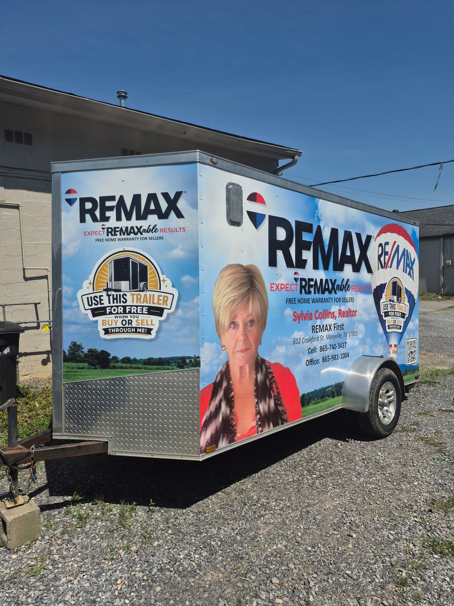 RE/MAX branded trailer parked on gravel beside a building under a blue sky