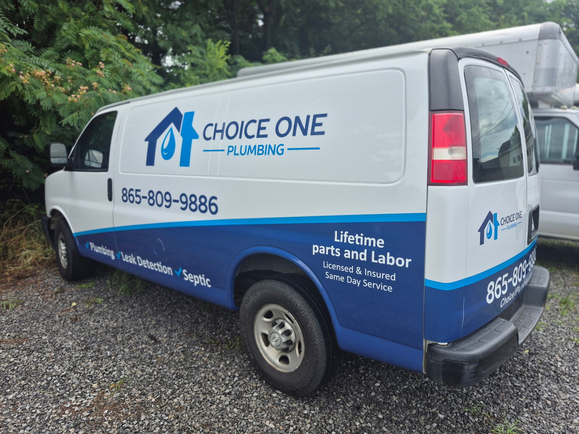 White and blue Choice One Plumbing van parked on gravel near trees and a fence