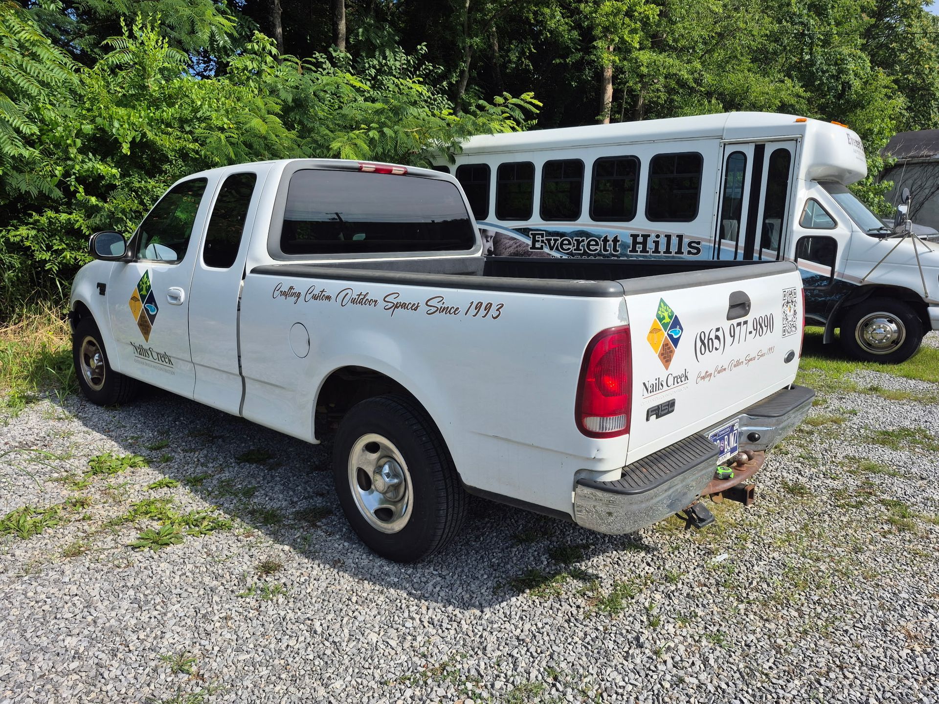 White utility pickup truck parked on gravel beside a white bus and trees