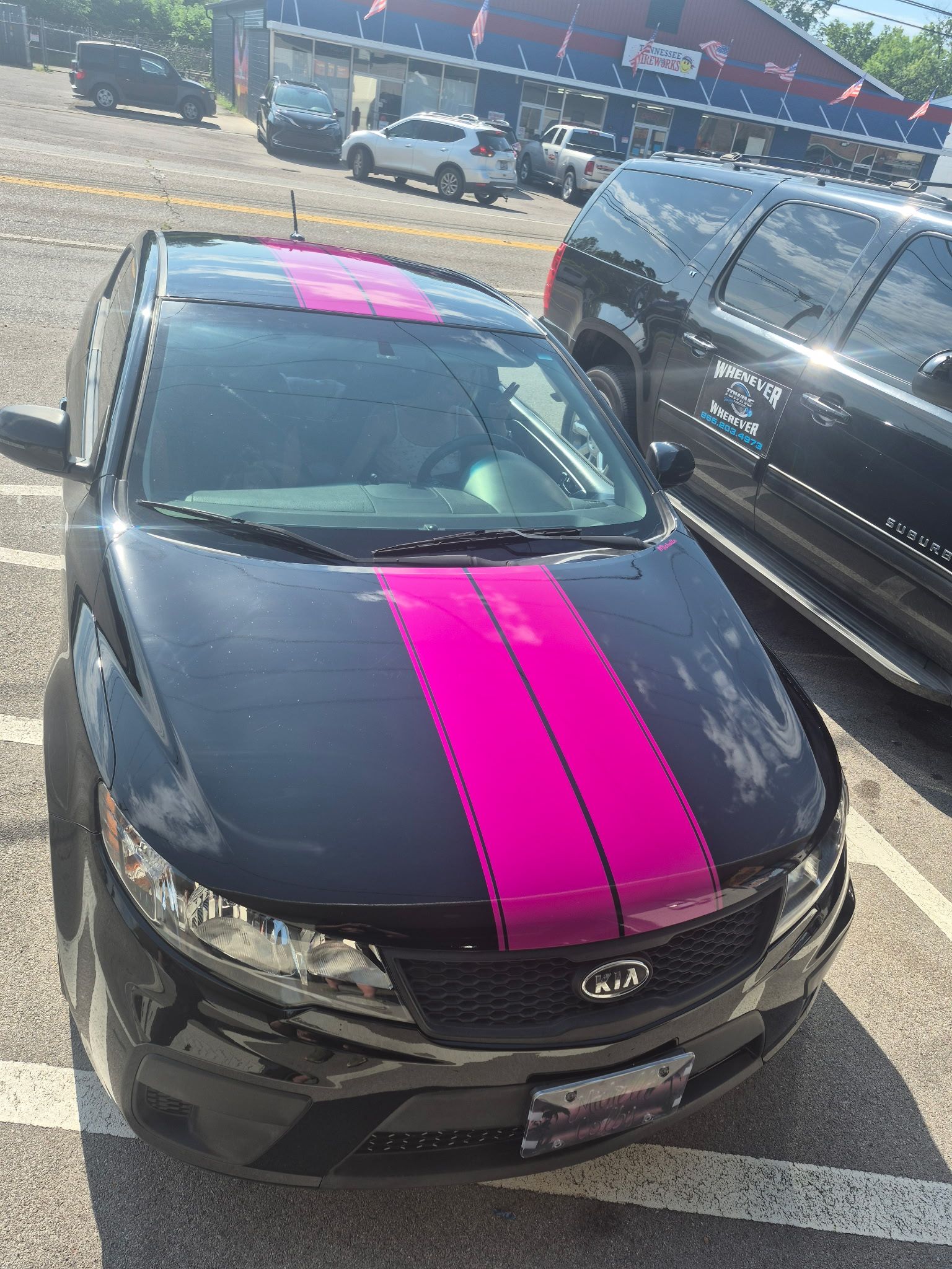 Black Kia sedan with bright pink racing stripes, parked in a lot beside a black SUV.