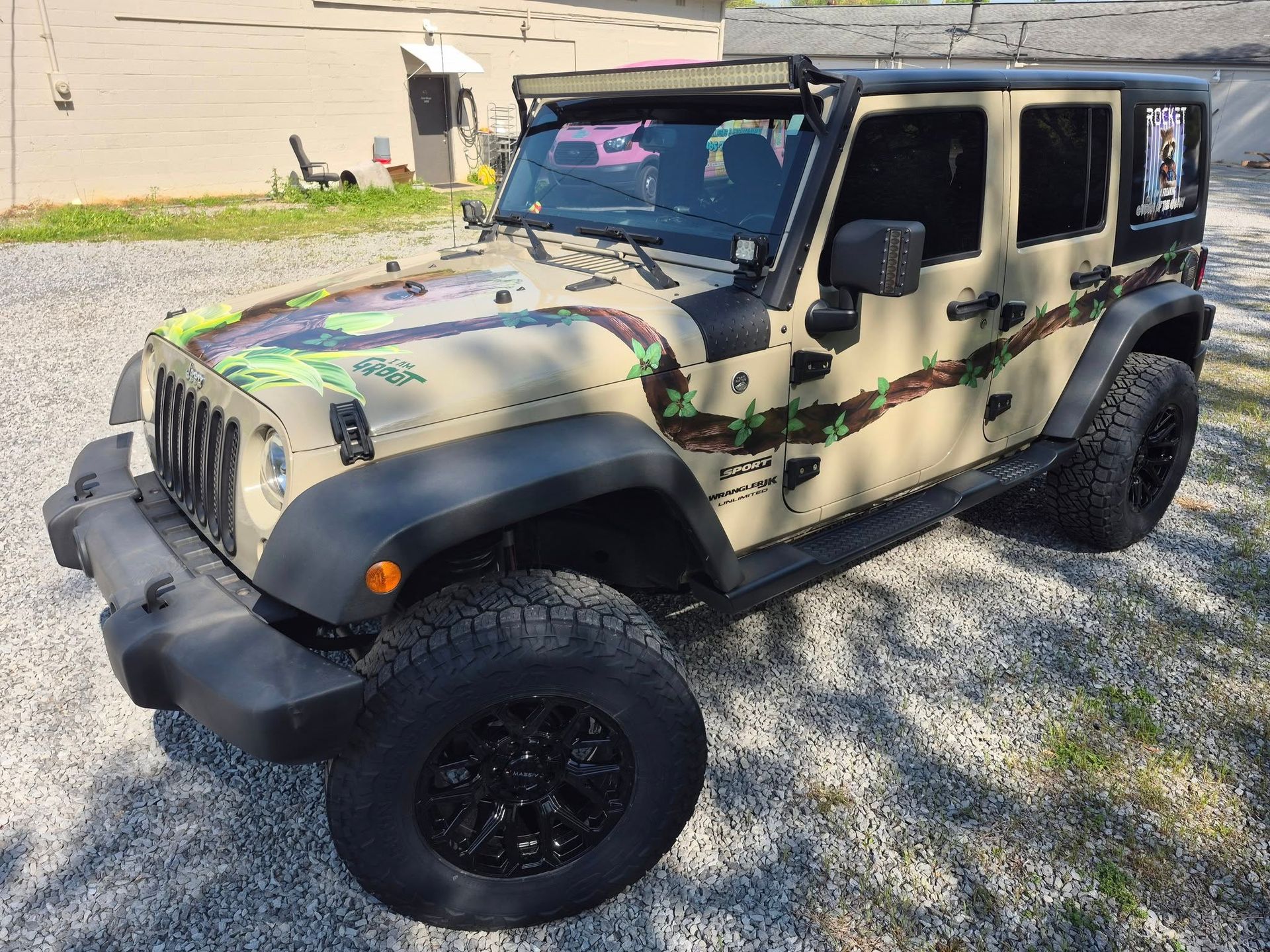 A jeep with a camouflage hood is parked in a gravel lot.