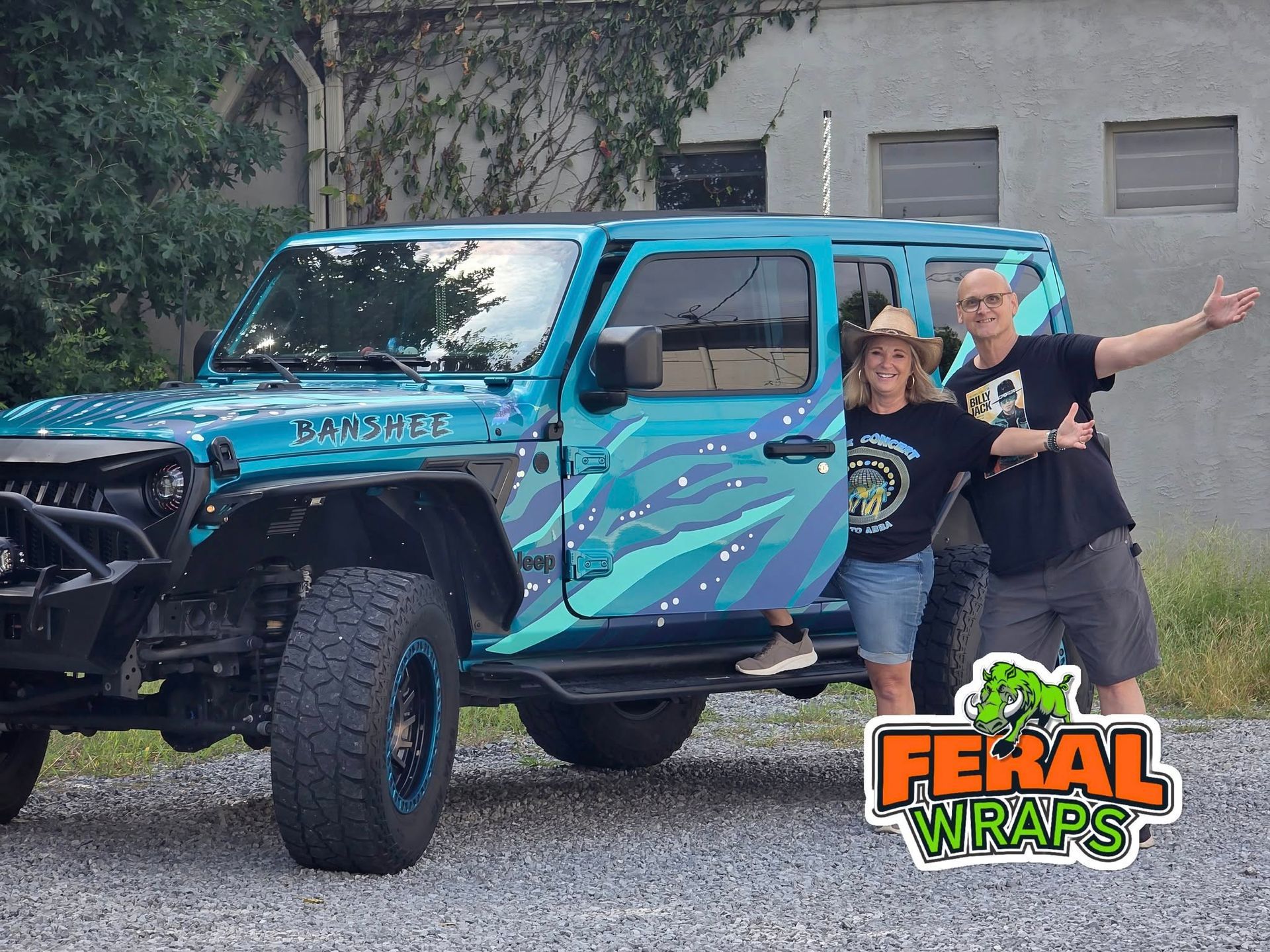 Couple poses by teal Jeep with graphic wrap. Man smiles, gives thumbs up. Outdoor setting, Feral Wraps logo.