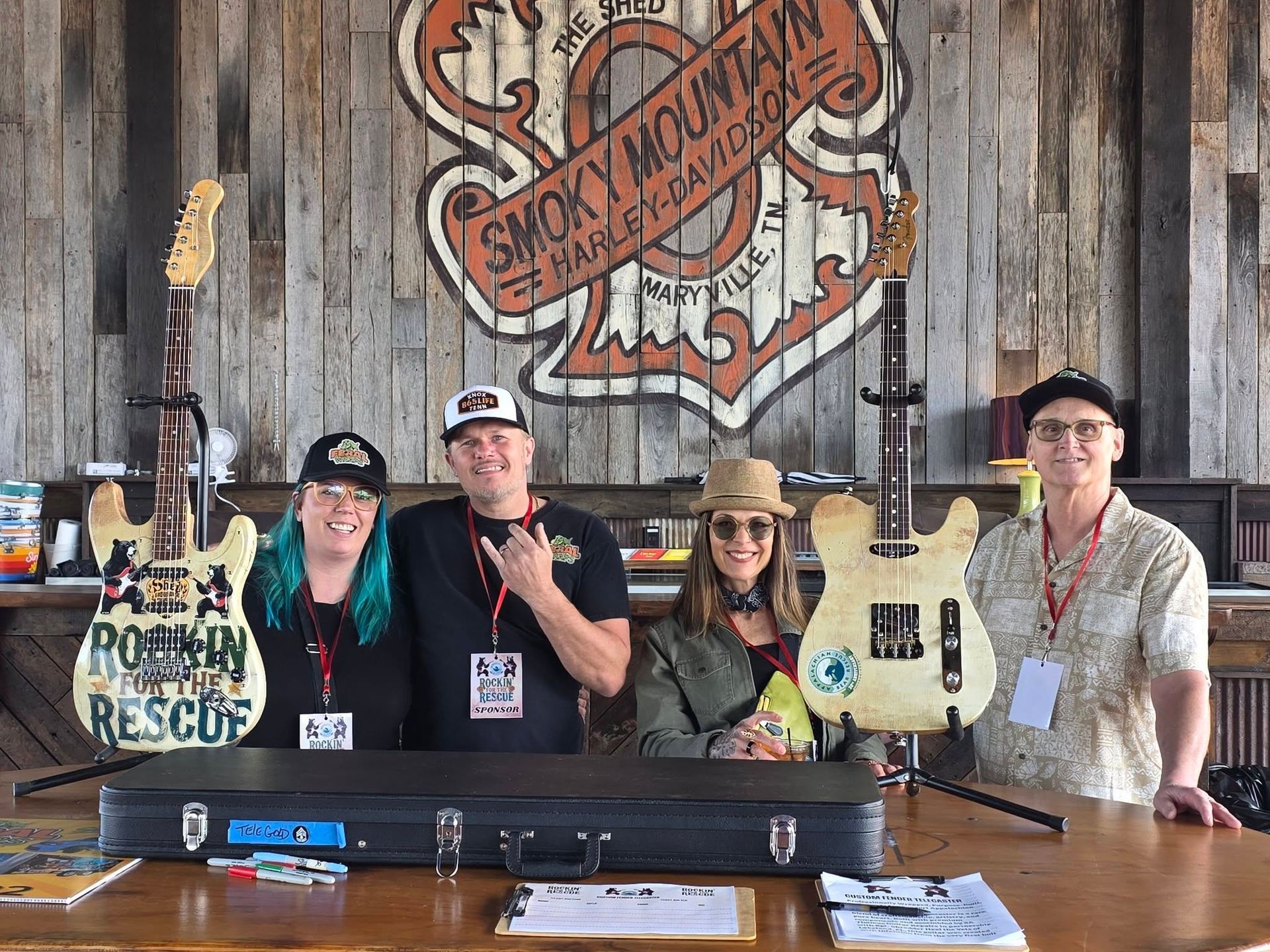 Four people posing behind a display table at Smoky Mountain Summerfest, with guitars and rustic wood backdrop