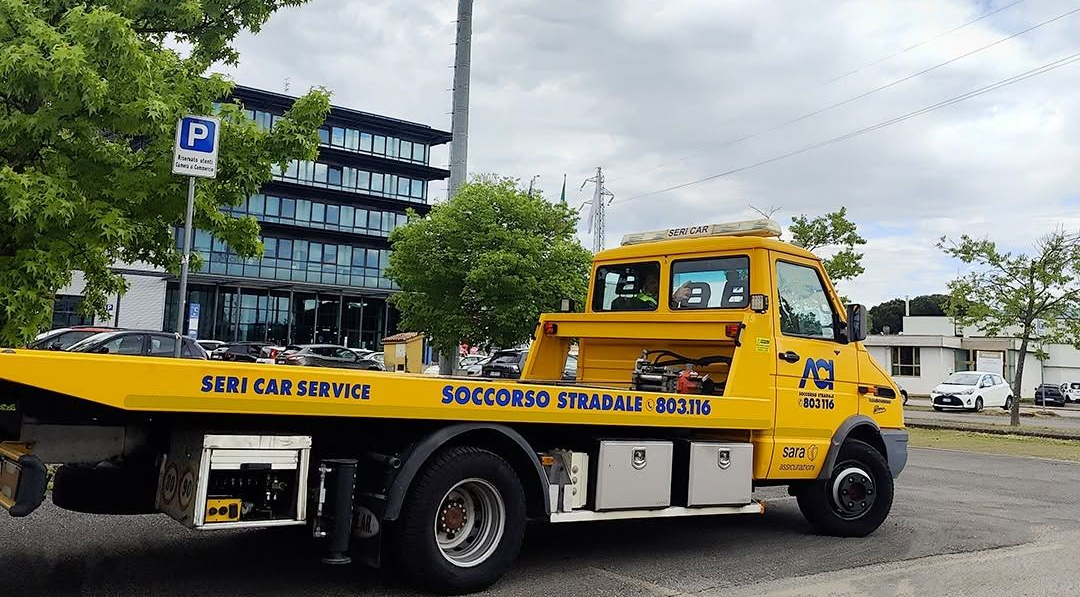 Carro attrezzi giallo parcheggiato su una strada accanto a un marciapiede e ad alcuni edifici, con la segnaletica di servizio sul lato.