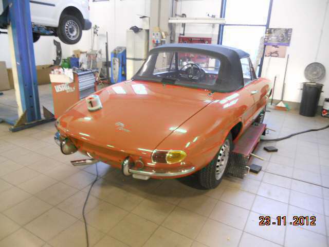 Una classica decappottabile arancione in un garage, circondata da attrezzi e da un'auto bianca sollevata.