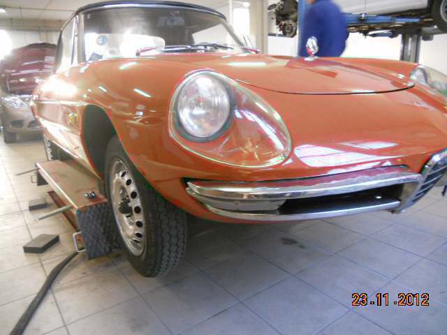 Auto sportiva d'epoca arancione su un ponte sollevatore in un garage, vista anteriore sinistra.
