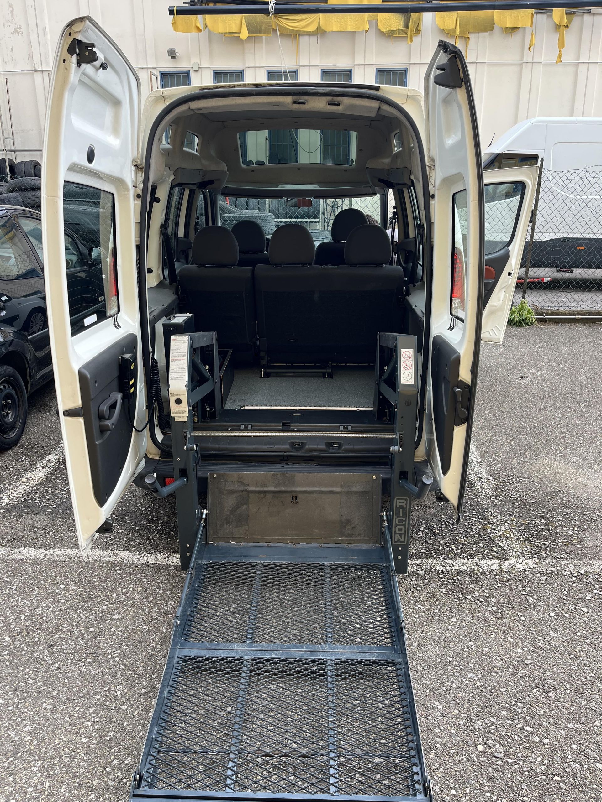 Furgone accessibile alle sedie a rotelle con rampa estesa, portelloni posteriori aperti, interni neri e sponde di sicurezza gialle.
