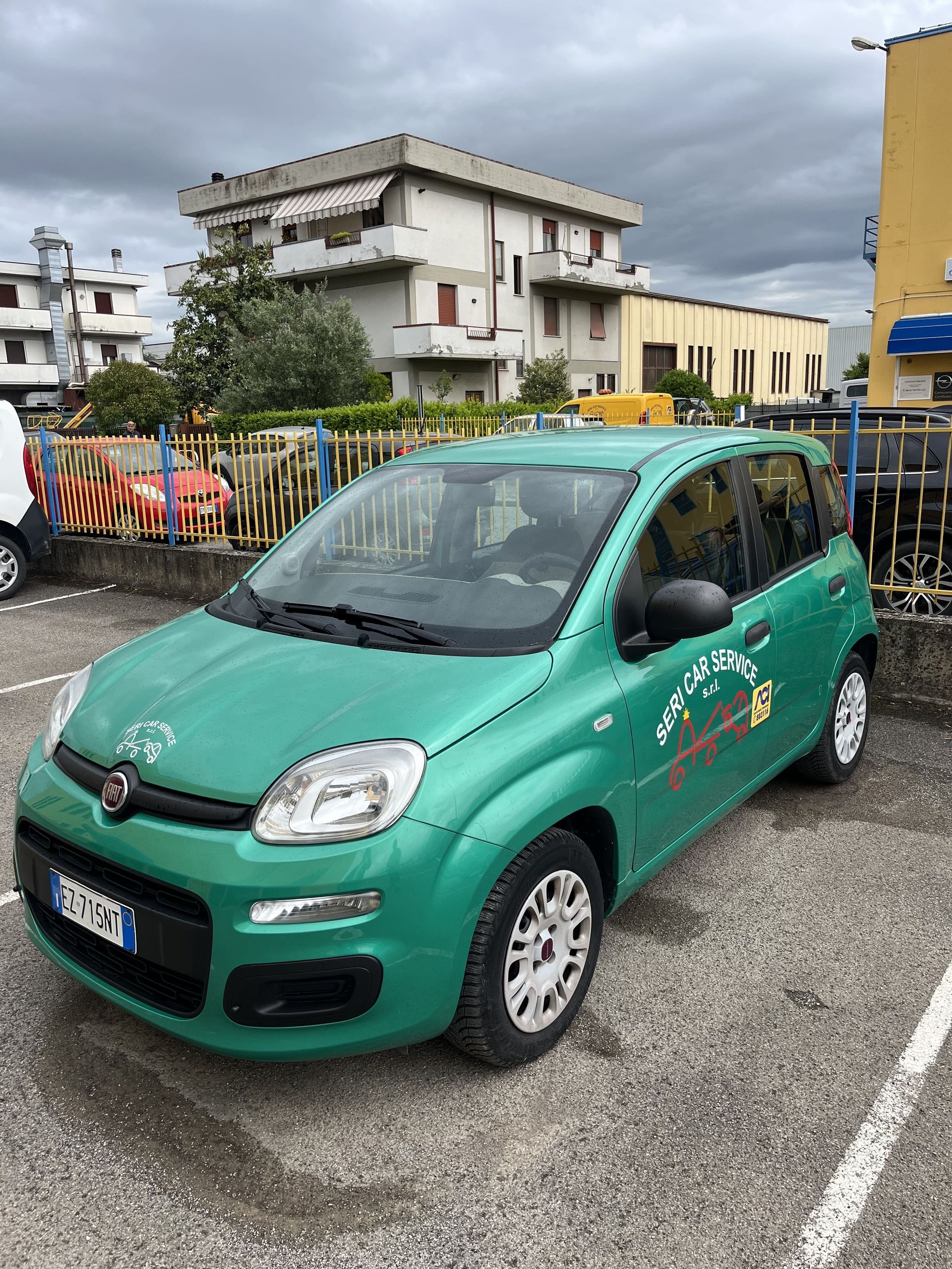 Un'auto compatta verde parcheggiata in un piazzale bagnato sotto un cielo nuvoloso, con edifici e una recinzione sullo sfondo.