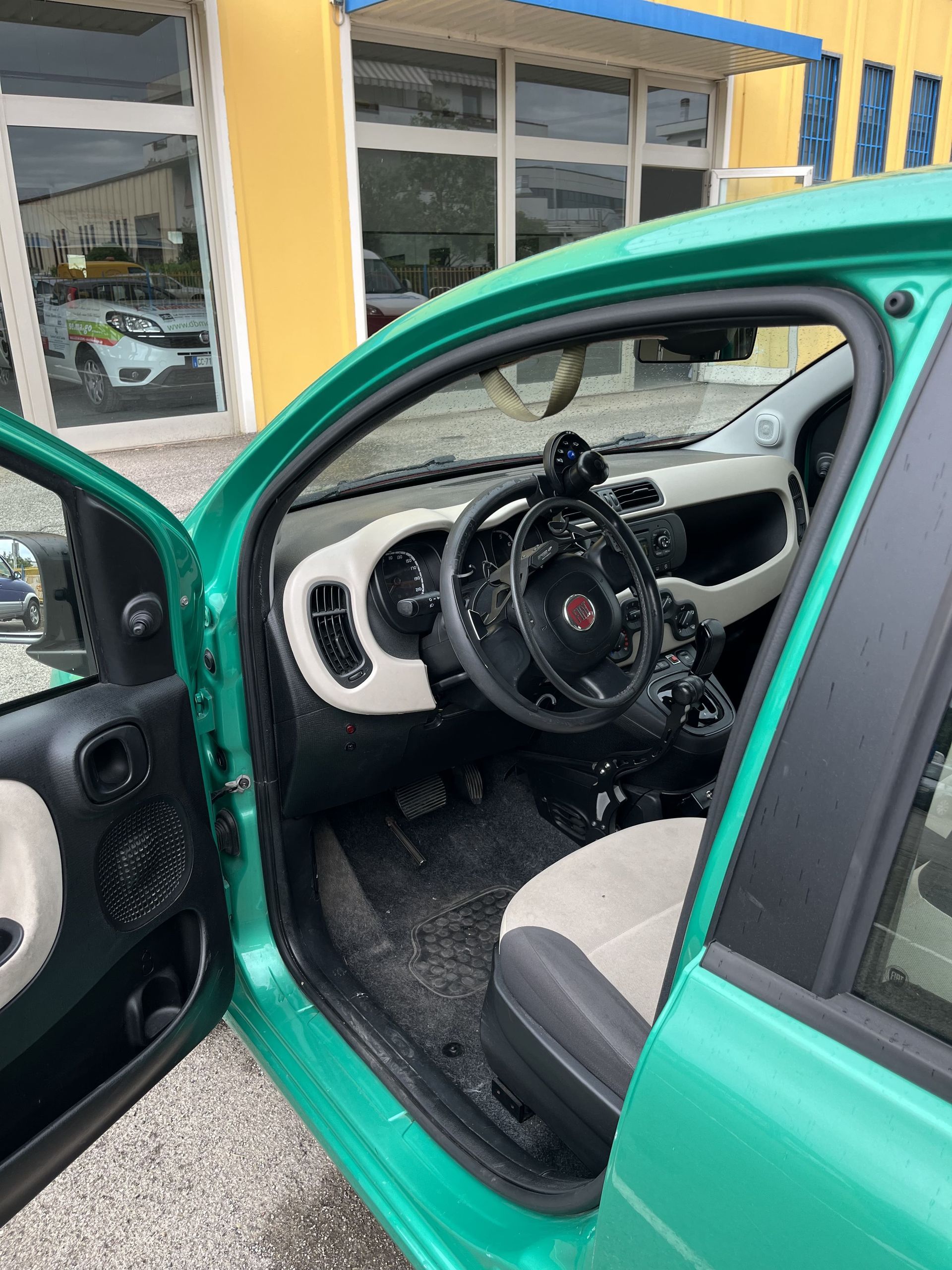 Un'auto verde con la portiera aperta, che mostra il volante e gli interni anteriori, parcheggiata davanti a un edificio.