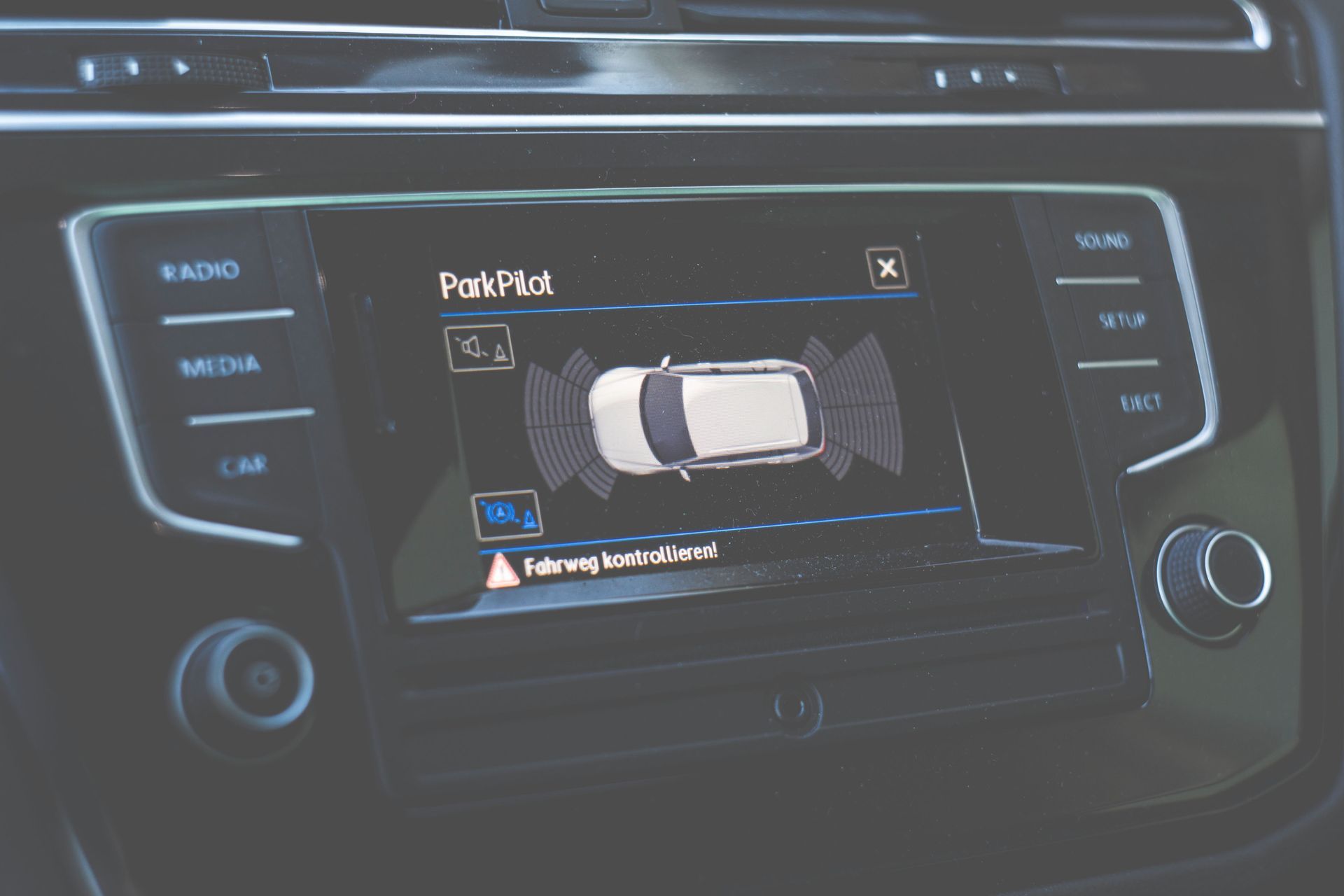Schermo dell'infotainment dell'auto che mostra l'assistenza al parcheggio con una visuale dall'alto dell'auto e archi dei sensori; comandi del cruscotto visibili