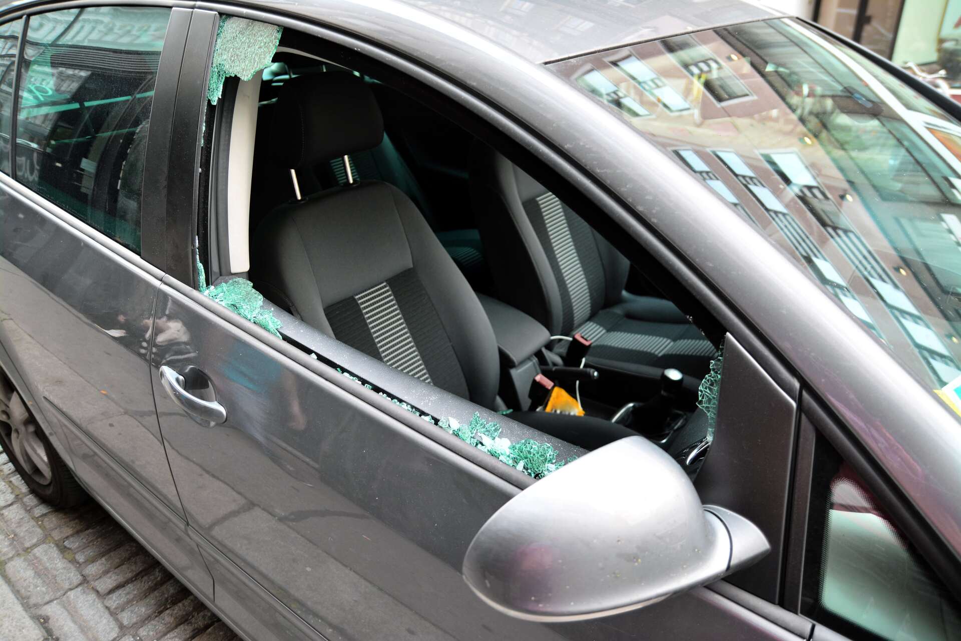 Auto argentata con parabrezza in frantumi e finestrino laterale rotto, detriti visibili all'interno del sedile anteriore.