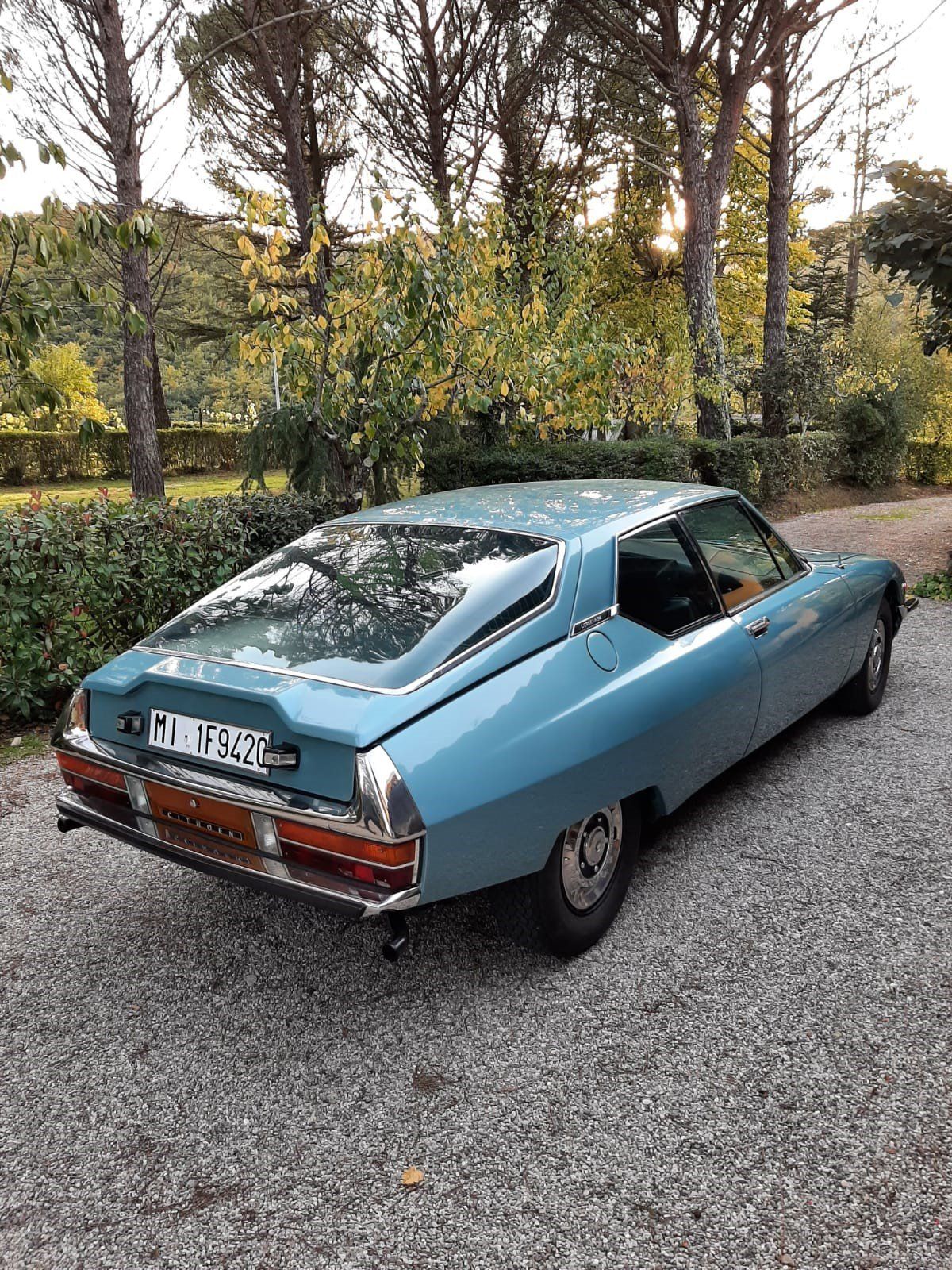Una coupé classica azzurra parcheggiata sulla ghiaia accanto a siepi e alberi.