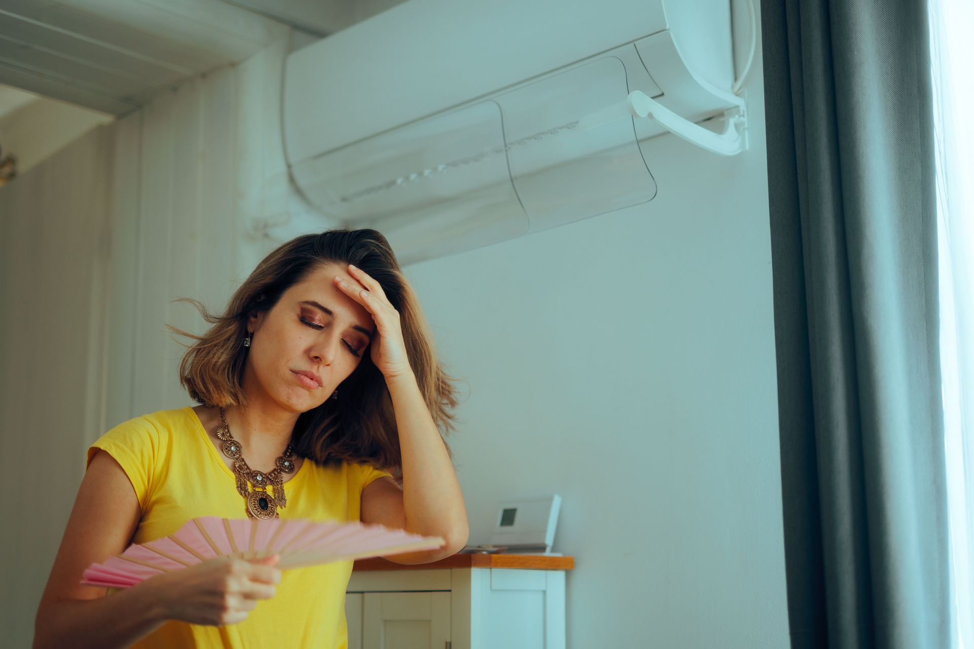 Woman looking overheated while fanning herself beneath a wall-mounted air conditioner. Woman looking overheated while fanning herself beneath a wall-mounted air conditioner.
