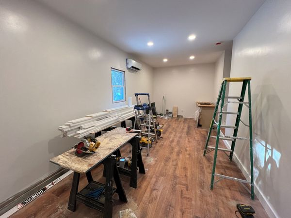 Narrow room under renovation with ladders, tools, worktable, wood flooring, and overhead lights.