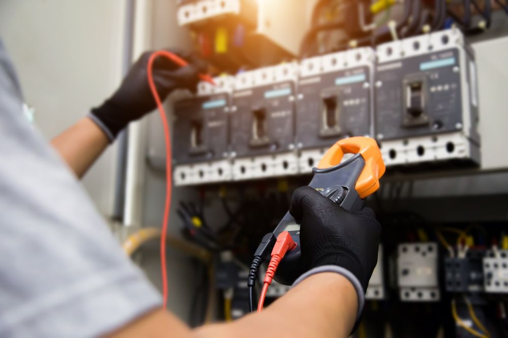 Electrician using a multimeter on an electrical control panel with exposed wiring.