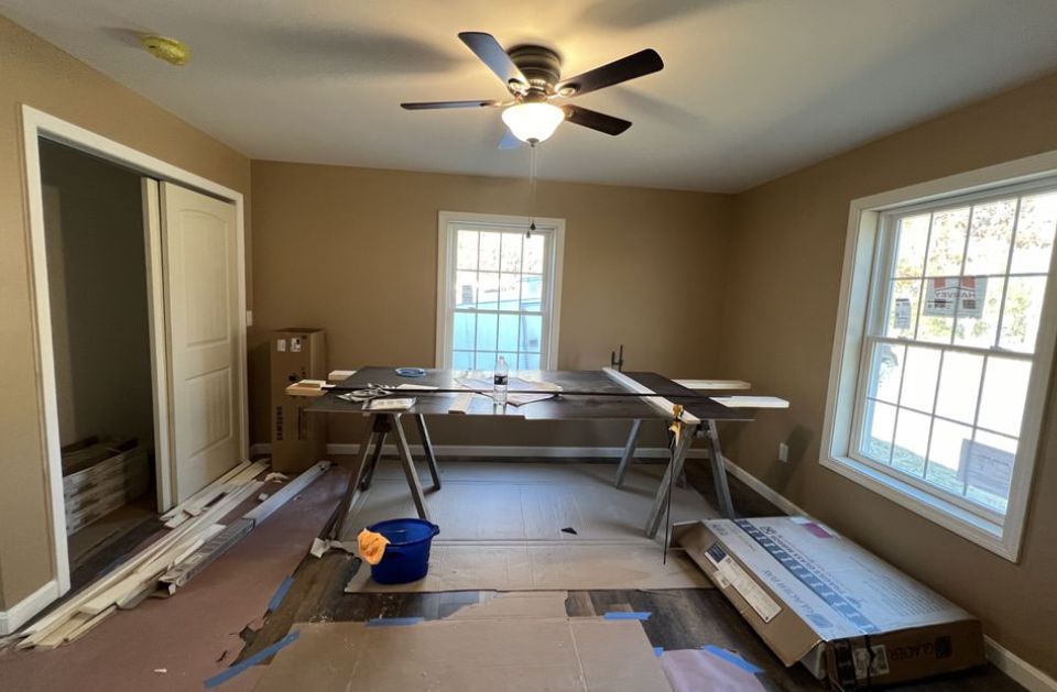 Unfinished room with painting supplies, drop cloths, ladder, ceiling fan, and bright windows.