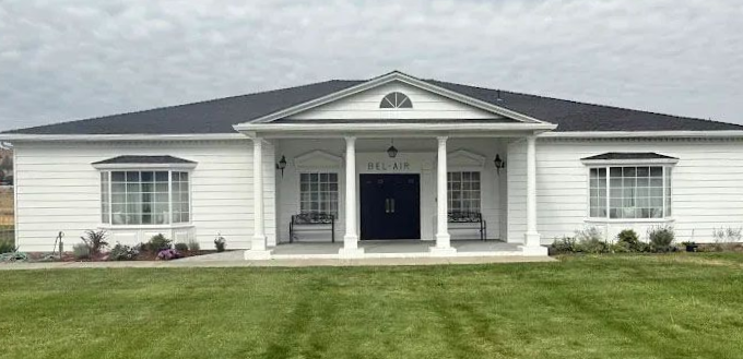 White house with black roof, columns, and bay windows, set on a green lawn against a cloudy sky.