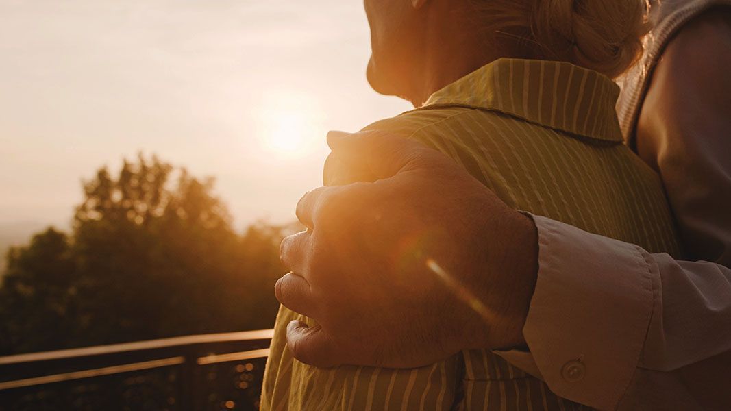 Person's arm around another, looking at sunset over trees.