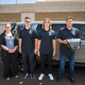 Four people in black shirts stand in front of a dark SUV, one holding a folded jacket.