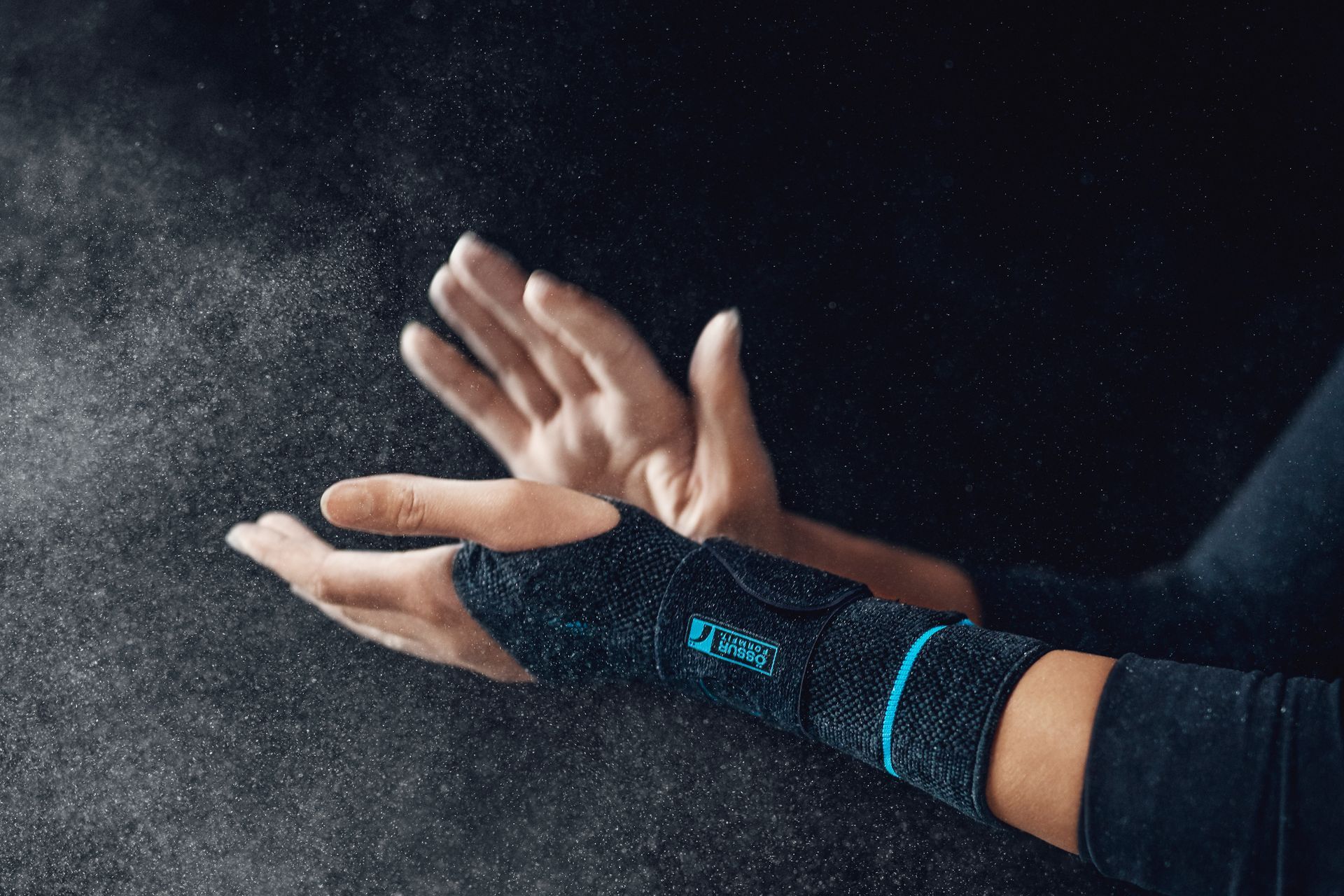 Hands clapping, releasing a cloud of chalk dust, with one hand wearing a black and blue Ossur wrist support brace.