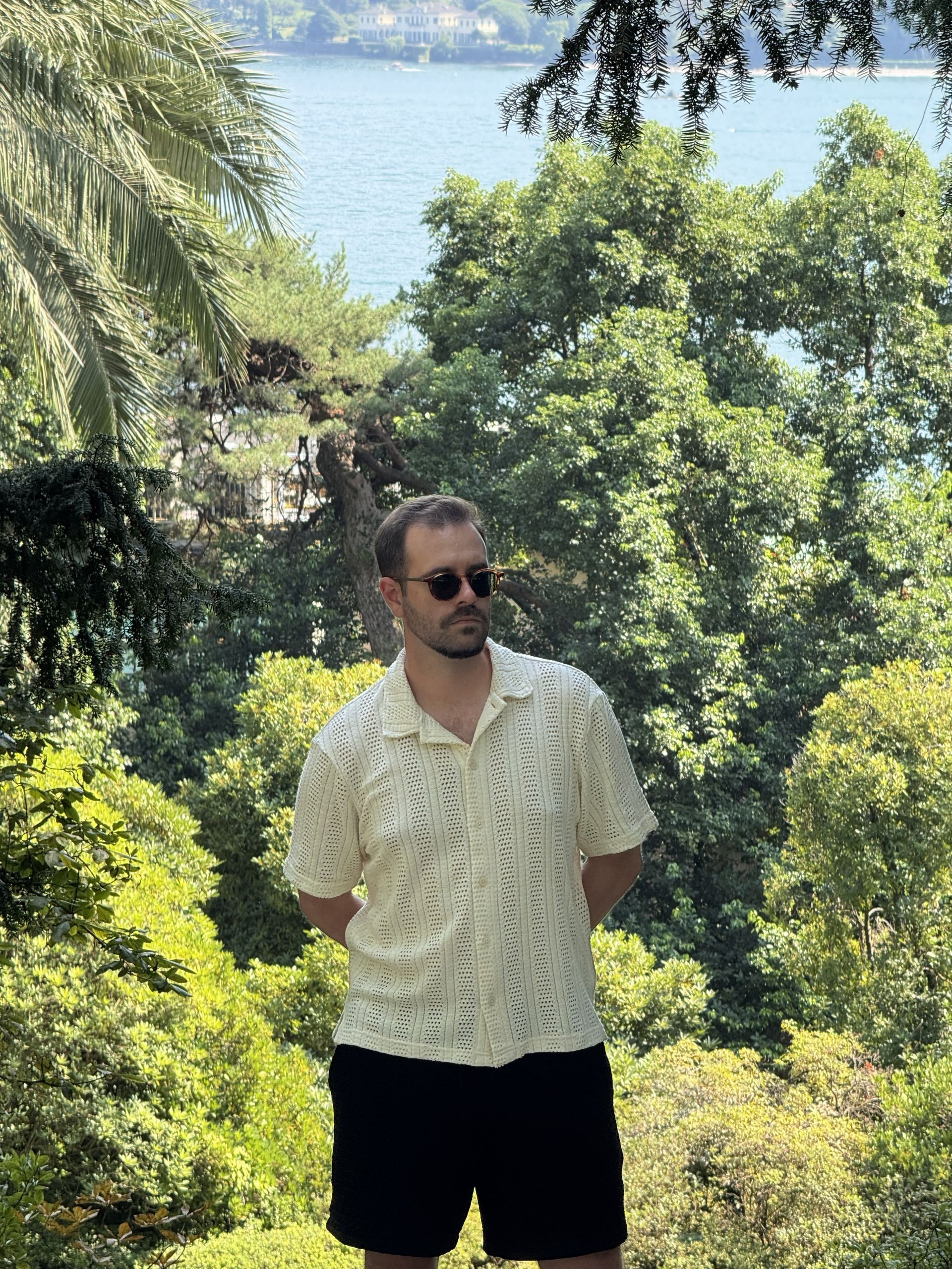 A person with sunglasses wears a patterned white shirt and black shorts, standing outdoors against a backdrop of trees.