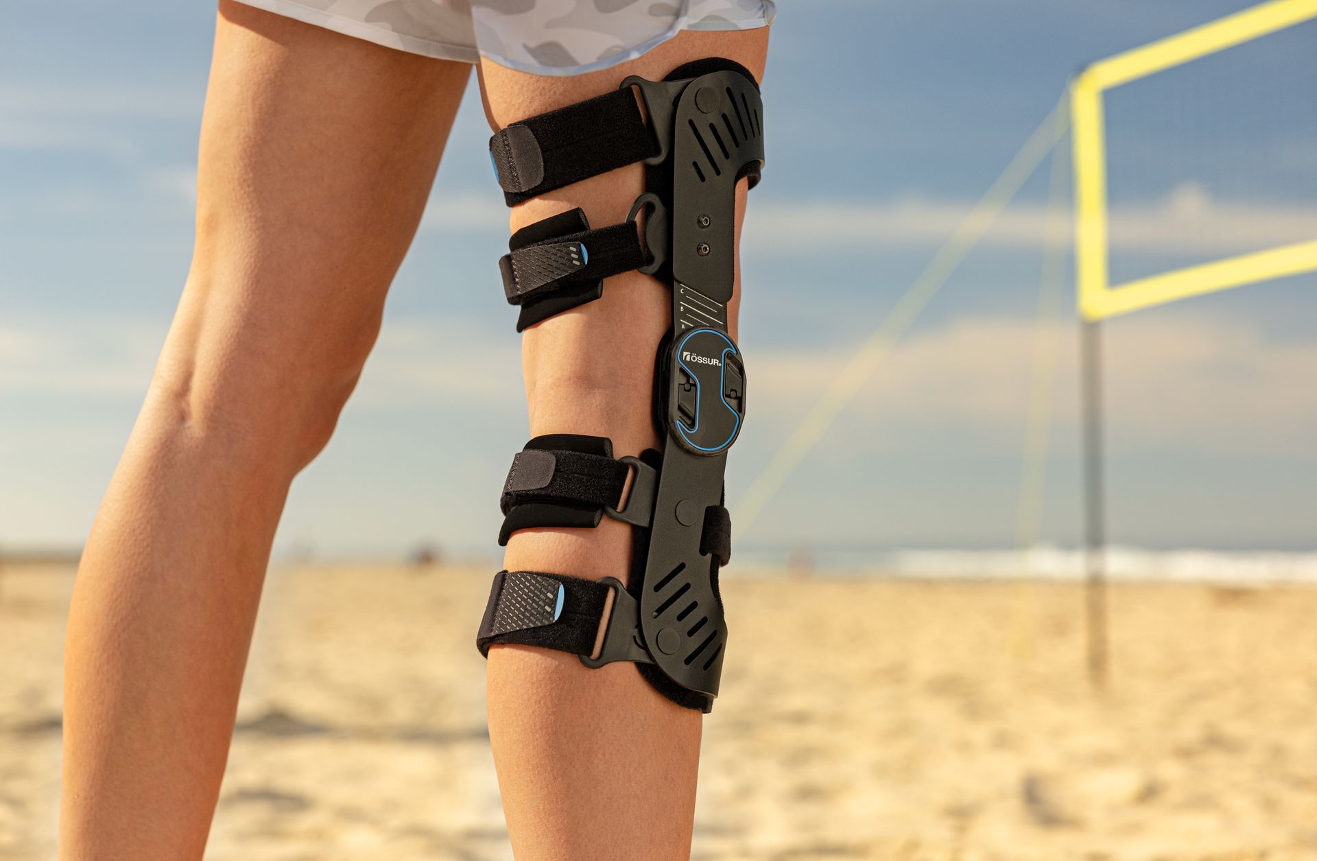 A person wearing a black knee brace on their leg while standing on a sandy beach near a volleyball net.