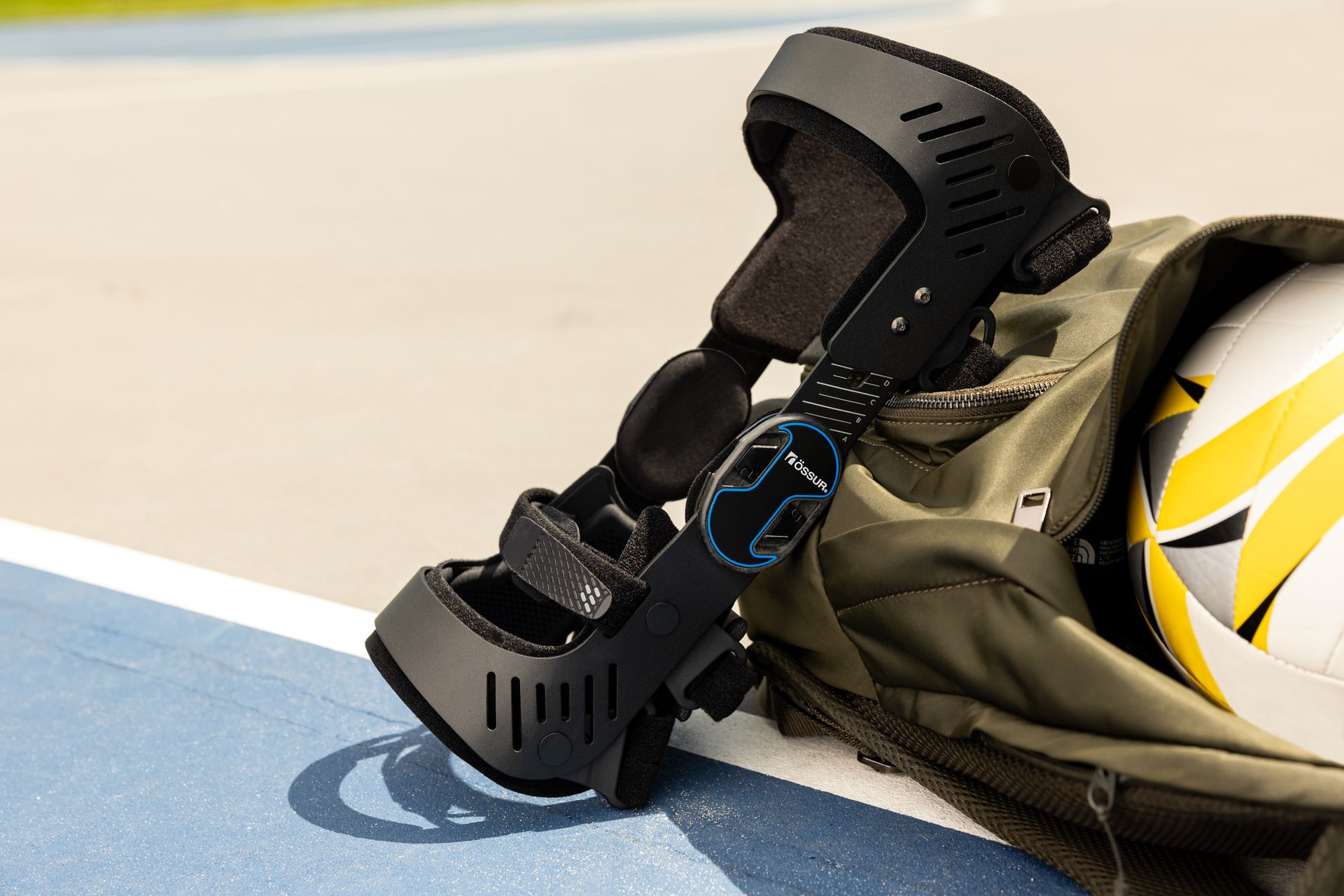 A black knee brace resting against an olive green backpack on a blue sports track.