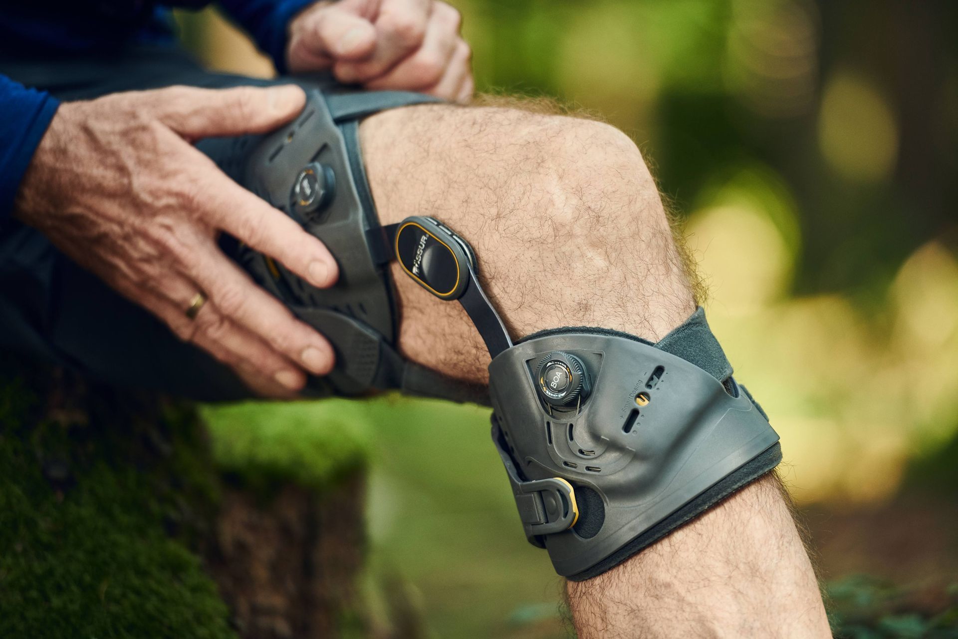 A person adjusts a grey knee brace on their leg, shown in an outdoor, wooded setting.