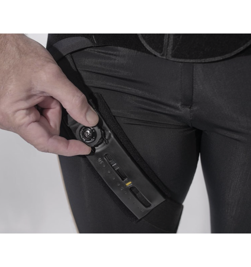 A hand adjusts a circular dial on a black mechanical brace or medical device attached to a person's thigh.