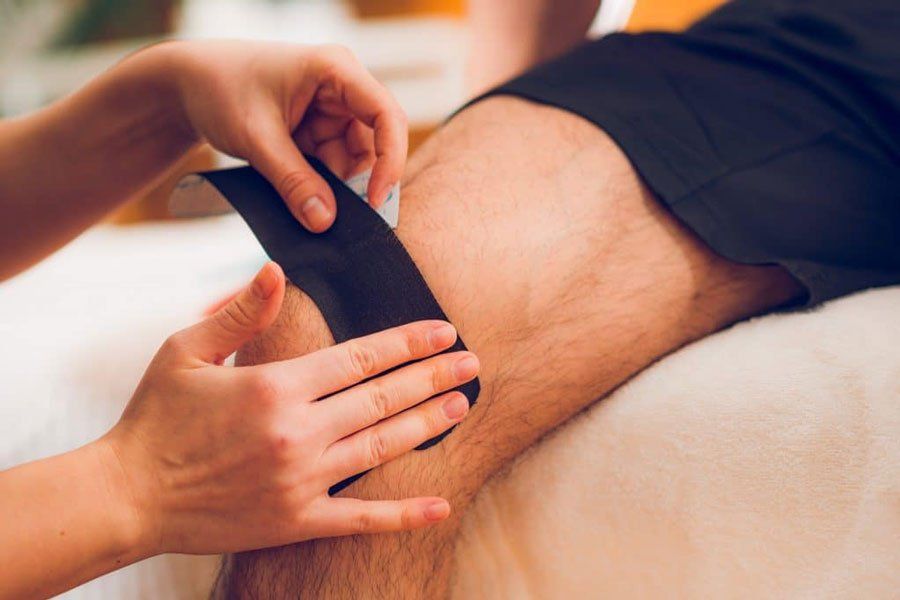 A person applies a black kinesiology tape strip to a knee in a clinical setting.