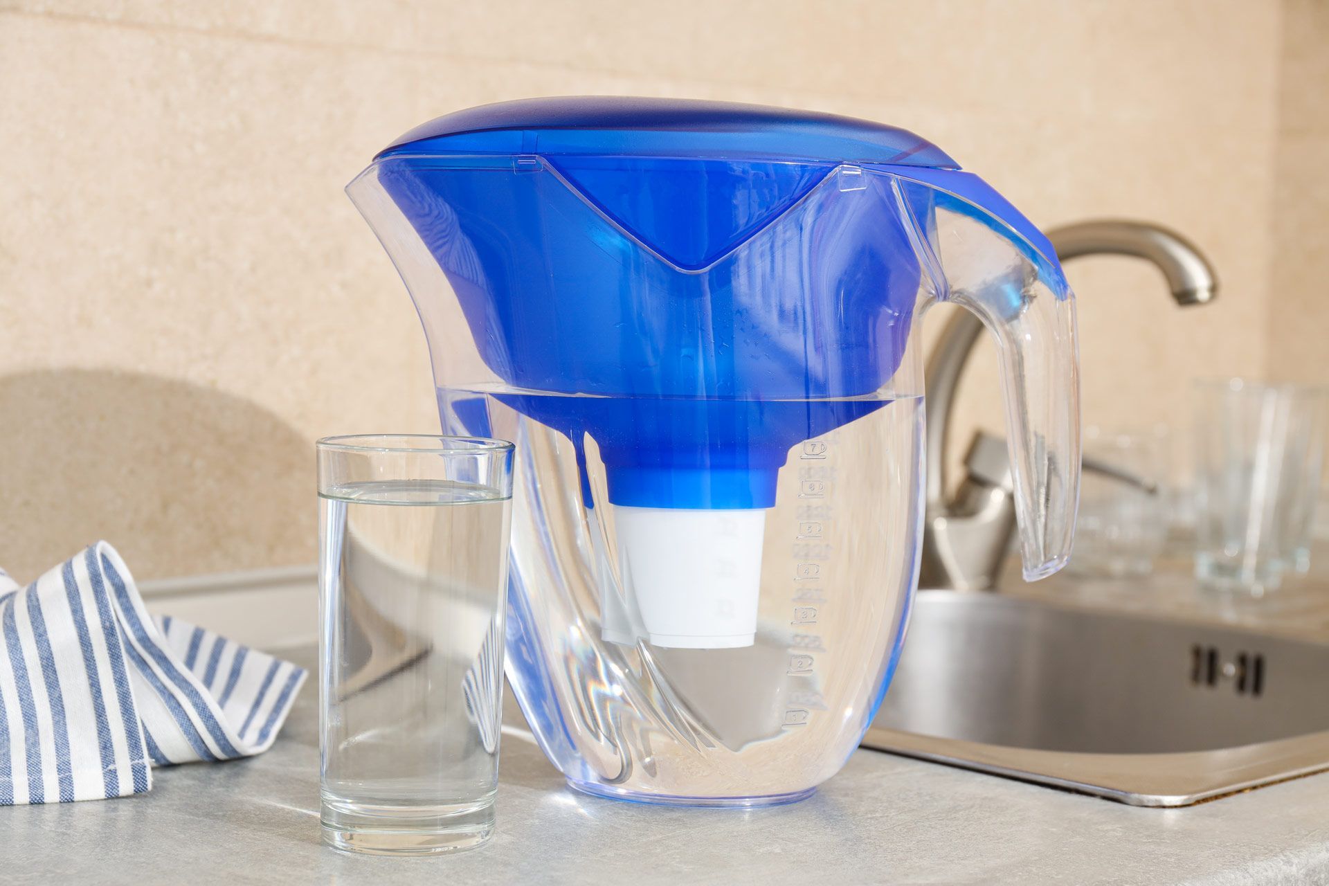 Blue water filter pitcher with a glass of water on a kitchen counter next to a sink.