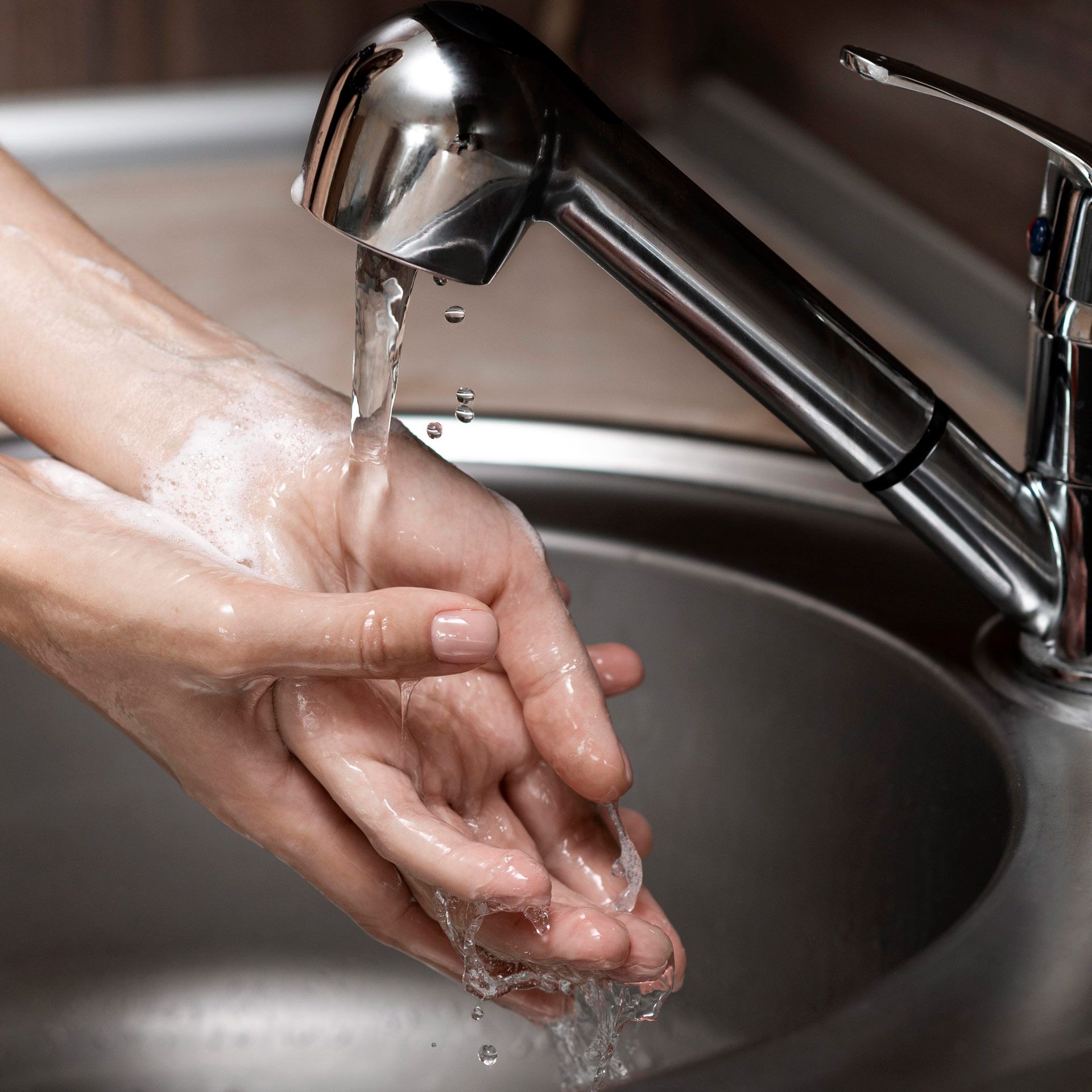Hands under running water in a stainless steel sink, rinsing suds from washing hands.