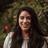 A woman with long dark hair is smiling for the camera in front of a tree.