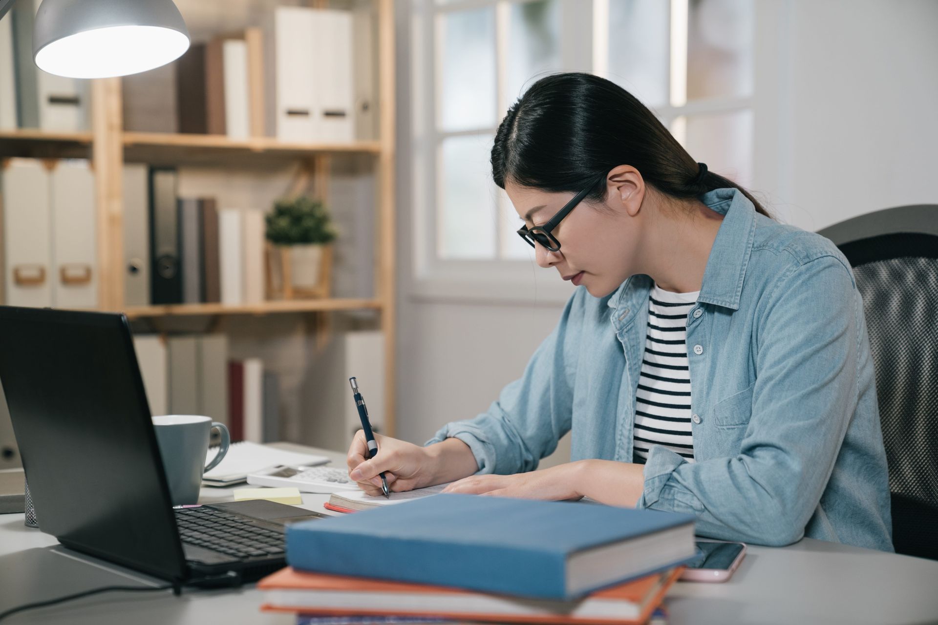 Person writing at a desk beside a laptop and stacked books in a bright office