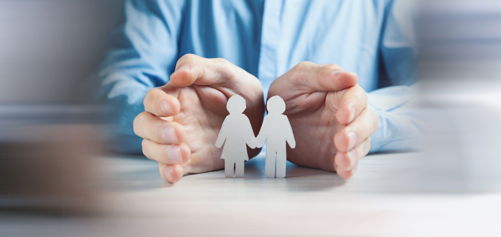 Hands shielding paper cutout family figures on a table, symbolizing protection and care