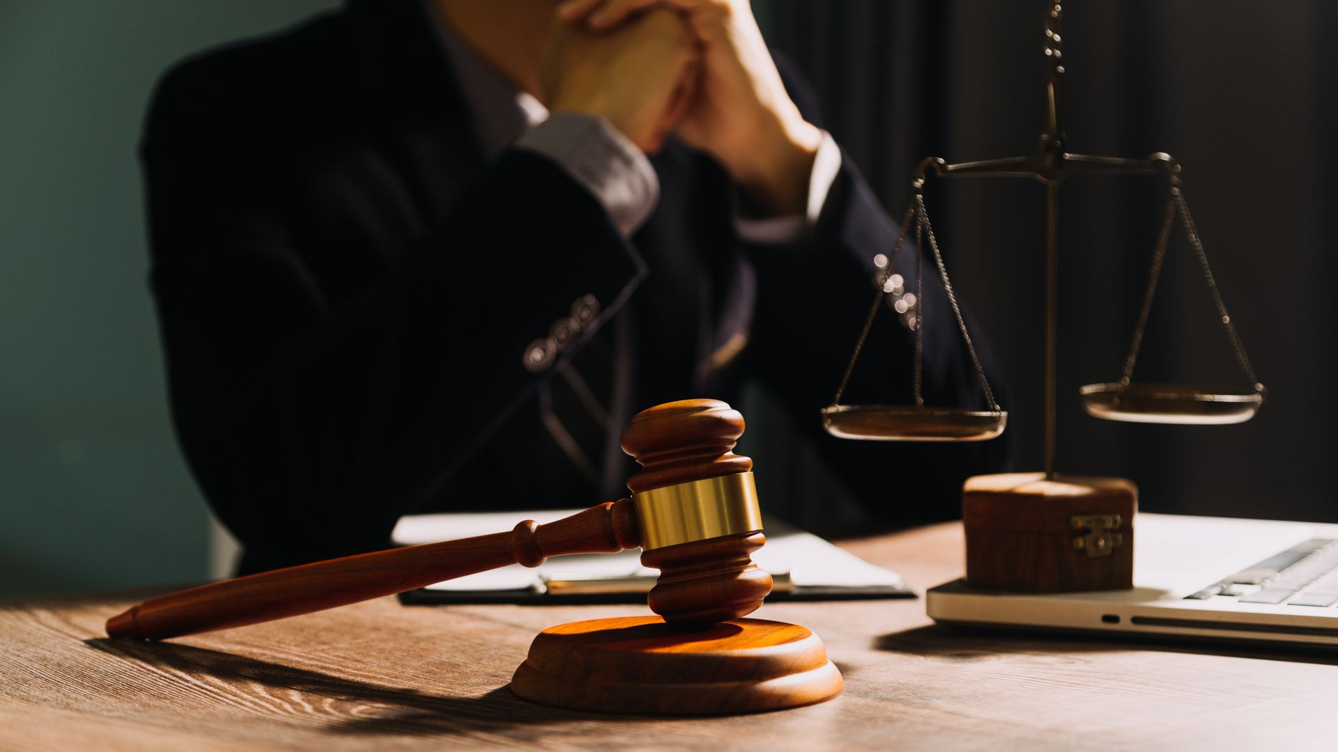 A legal gavel and scales of justice on a desk in front of a person in a suit, with dark, professional lighting.