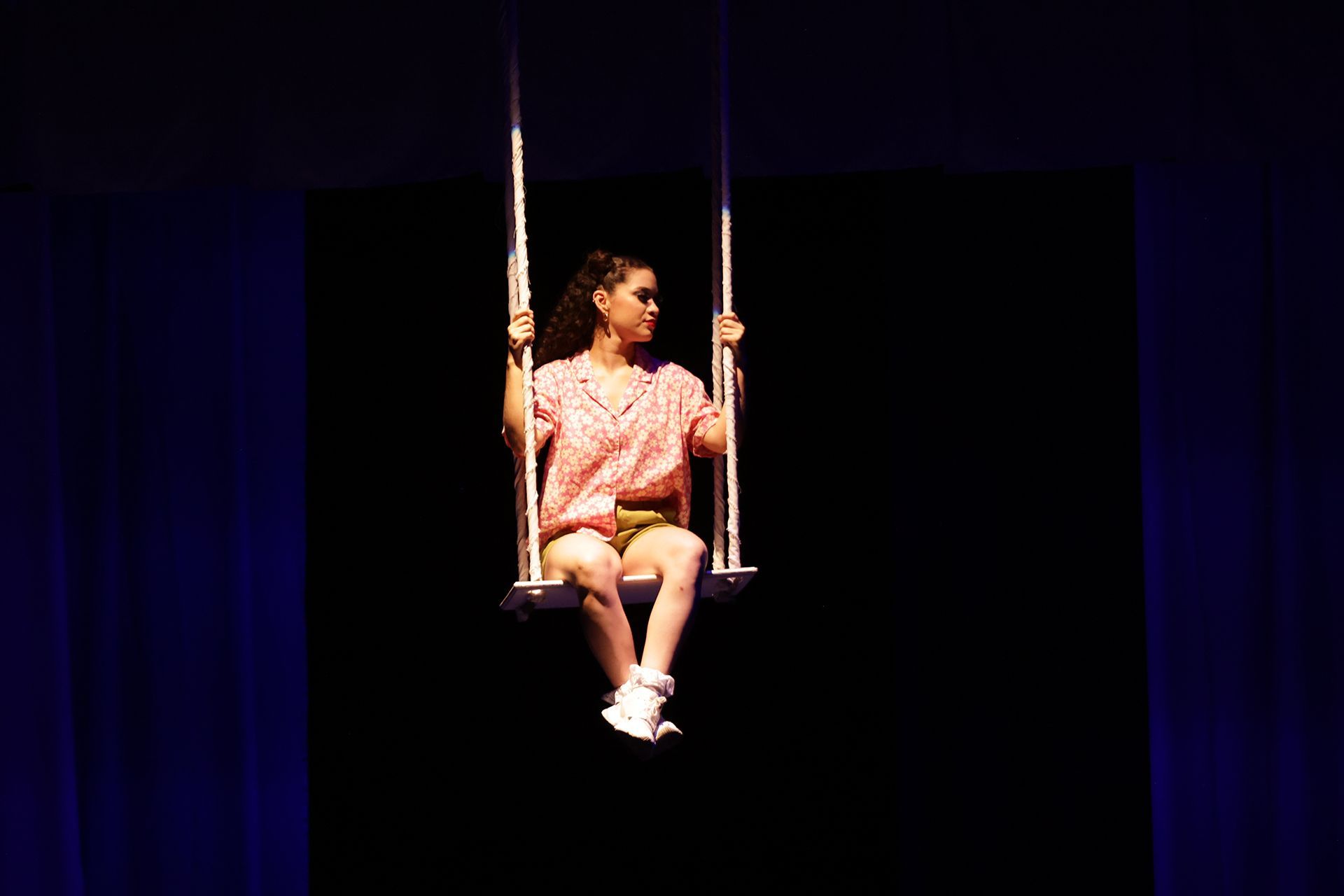 Mujer en un columpio con fondo negro, vestida con una camisa rosa, pantalones cortos y zapatos blancos.