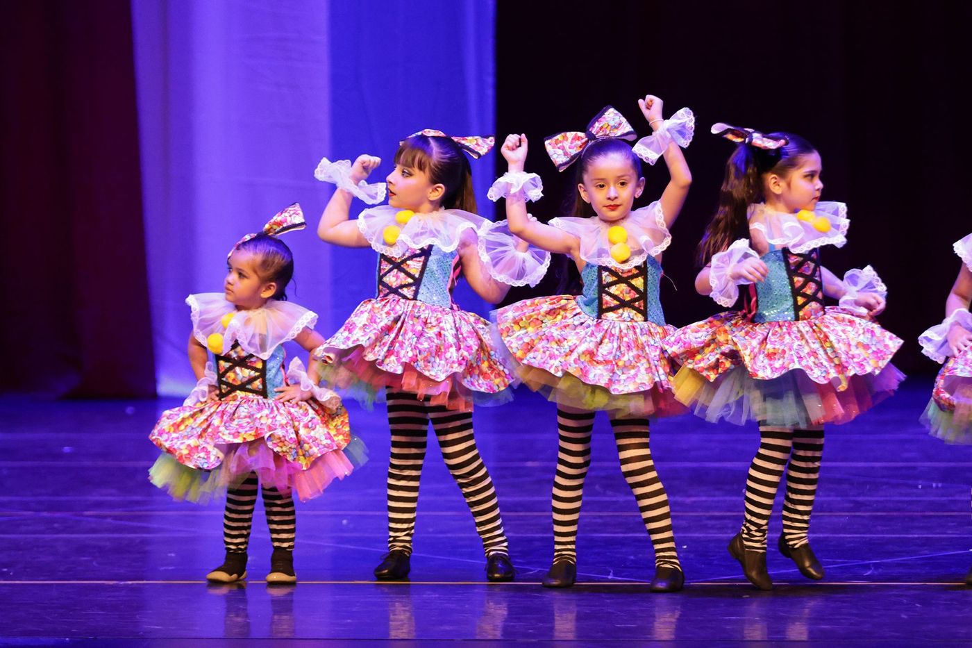 Cuatro jóvenes bailarinas con coloridos trajes y mallas a rayas en el escenario, con los brazos en alto.