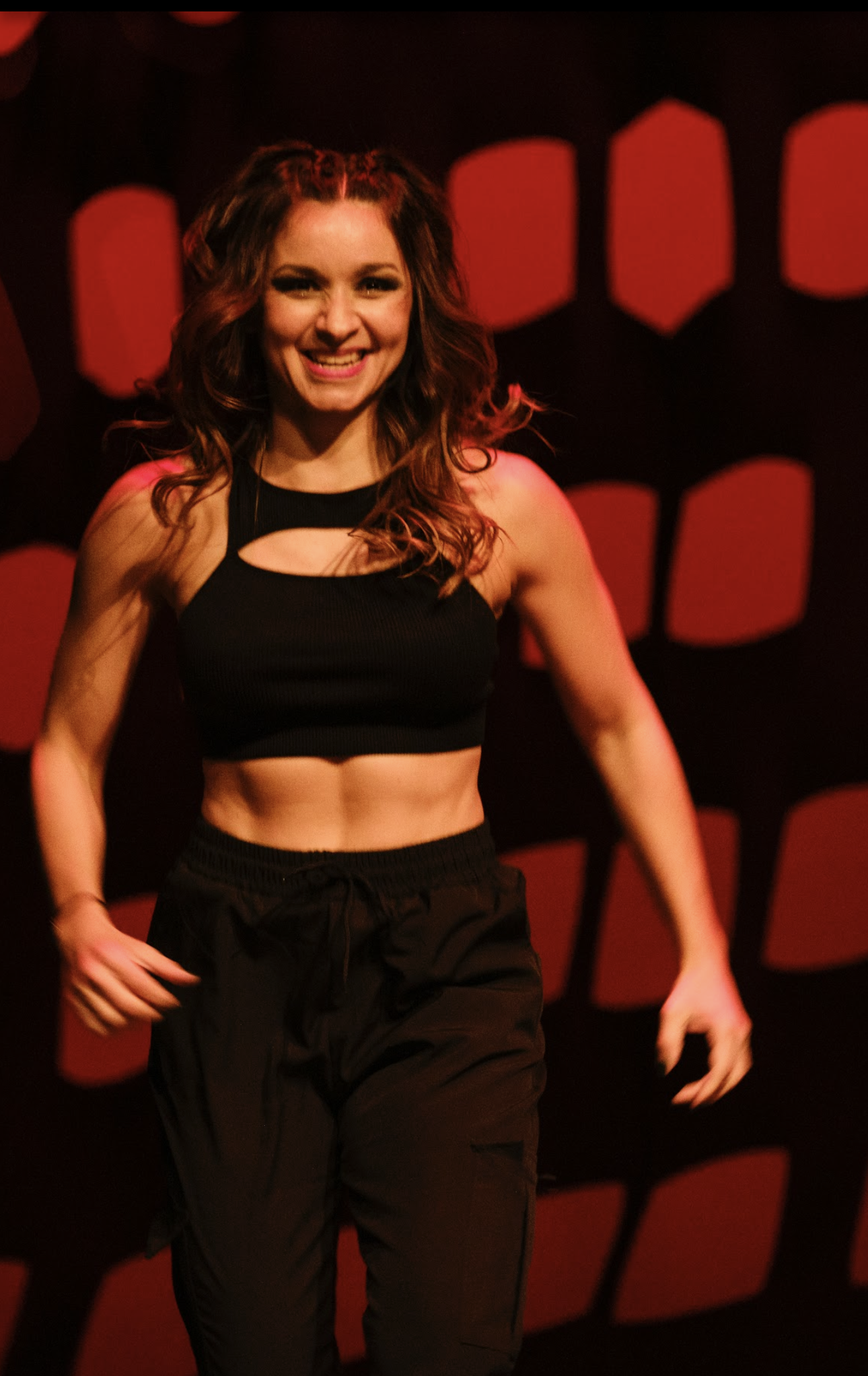 Mujer con top corto y pantalones negros sonríe en el escenario con fondo rojo.