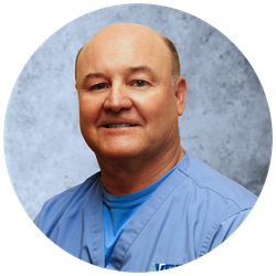 A bald man in a blue scrub top smiles for the camera